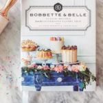 Bobbette & Belle Classic Recipes cookbook displayed on a marble surface with baking confetti and a rolling pin