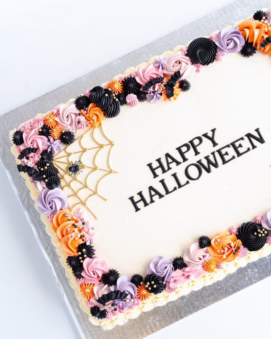 Halloween Slab Cake 1 A rectangular sheet cake decorated for Halloween, with a smooth white icing base and a colorful buttercream border. The border features intricate piping in shades of orange, black, purple, and pink, forming a festive and playful design. On one corner, a gold spider web is piped delicately, complete with a small decorative spider. The center of the cake displays the words "Happy Halloween" in a bold, elegant font, creating a clean and stylish look. The cake sits on a silver cake board, making it a beautiful centerpiece for any Halloween gathering.