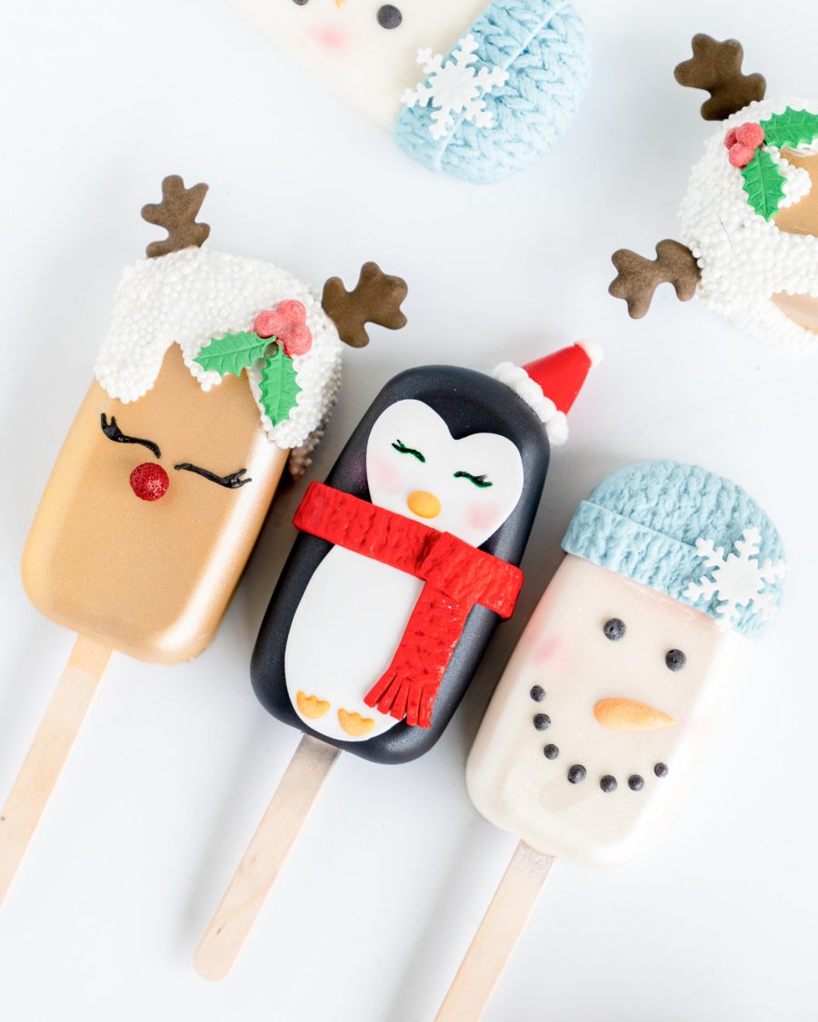 Christmas Cake pops 1-1 Holiday-themed cake popsicles featuring hand-decorated designs of a reindeer with holly accents, a penguin with a red scarf and Santa hat, and a snowman with a blue knitted hat and snowflake decoration.