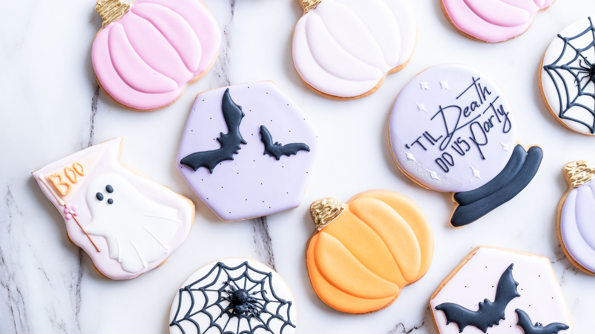 An assortment of Halloween-themed sugar cookies featuring designs of pastel pumpkins, ghosts, bats, spiderwebs, and a crystal ball with a festive message.