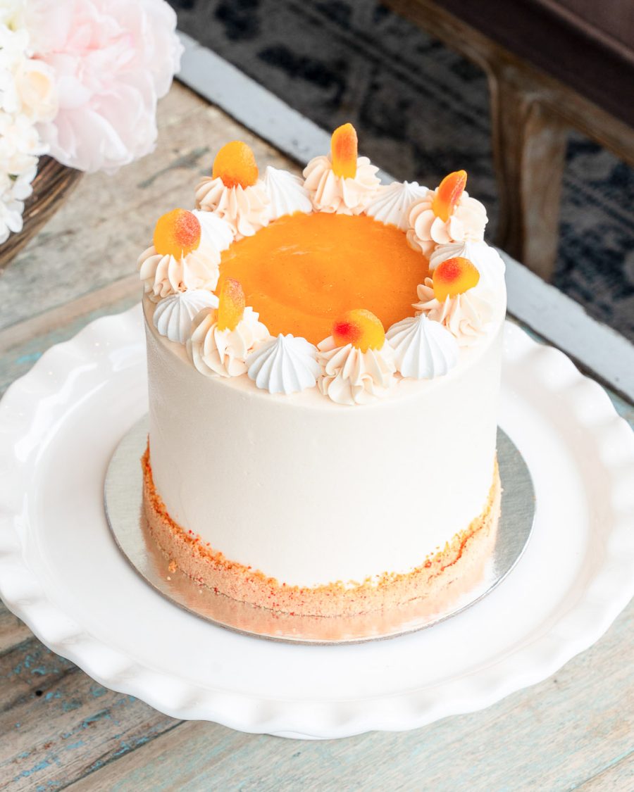 Peaches & Cream Cake Peaches and Cream Cake topped with peach purée, meringues, buttercream swirls, and fuzzy peach candy on a white plate.