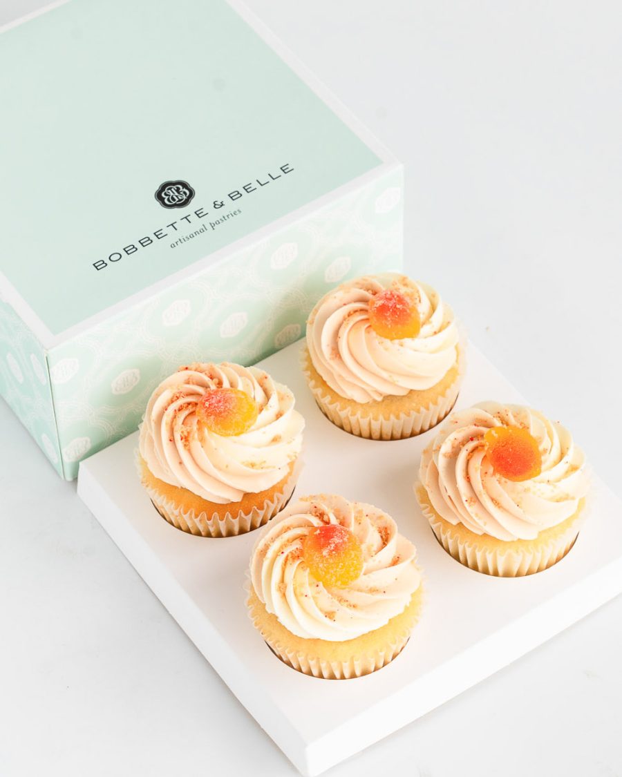 Four vanilla cupcakes topped with peach buttercream and fuzzy peach candies in a pastel bakery box.