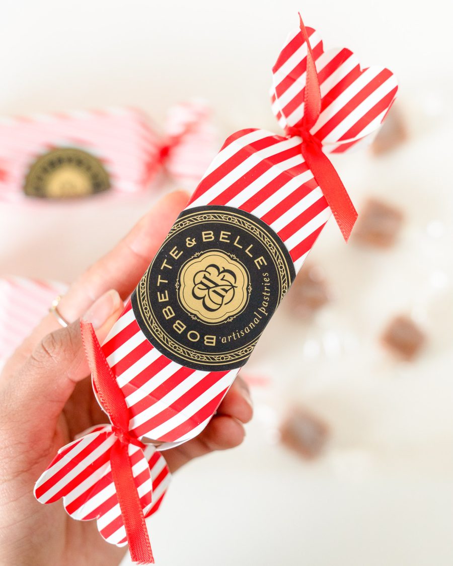 Hand holding a red-and-white striped Bobbette & Belle holiday caramel cracker with gold and black artisanal label.