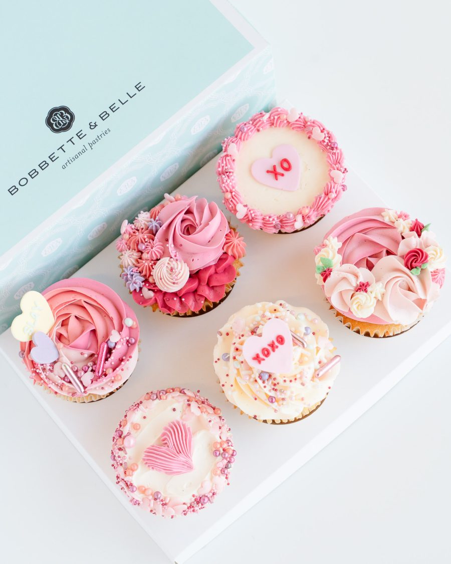 6 piece Valentines Cupcakes A gift box of four Valentine’s cupcakes decorated with pink, red, and cream buttercream piping and heart toppers.